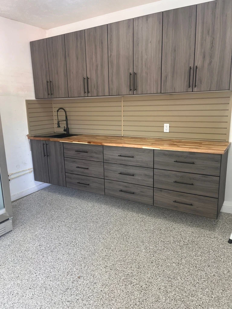 A high-end storage setup illustrating how a Garage Makeover Value to Your Home increases, featuring wood-grain textured floating cabinets, a butcher block countertop with a built-in sink and matte black faucet, tan slatwall backsplash, and a premium grey epoxy floor.