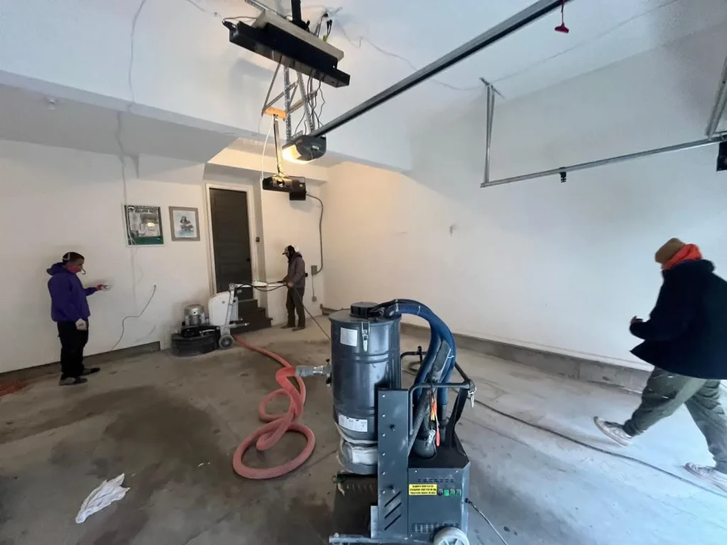 A crew of professionals preparing a concrete floor, illustrating How Long Does a Garage Makeover Take, using industrial grinders and a heavy-duty vacuum system to ensure a clean surface for a high-quality epoxy coating.