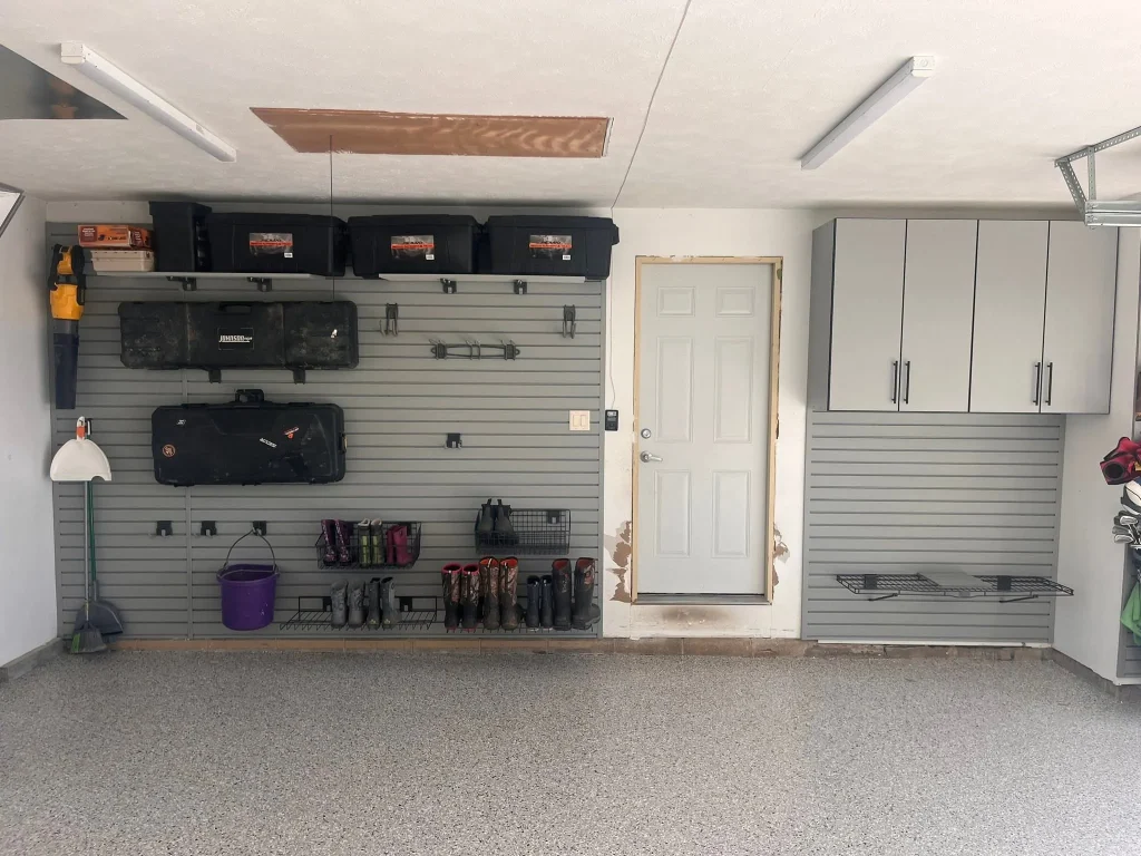 An organized interior highlighting What Does a Garage Makeover Actually Include, featuring grey slatwall panels with overhead storage bins, wall-mounted boot racks, gear cases, and light grey upper cabinets above a clean epoxy floor.
